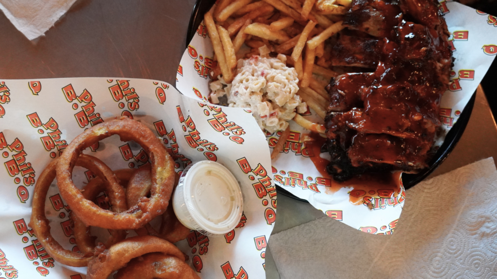 聖地牙哥 Phil's BBQ 餐廳內餐桌上的當天點餐全景特寫。畫面上擺放著金屬托盤盛裝的豐盛美式烤肉套餐，包含塗滿招牌甜醬的 Baby Back Ribs（豬肋排）、現炸黃金薯條（French Fries）、新鮮高麗菜沙拉（Coleslaw）與通心粉沙拉（Macaroni Salad）。