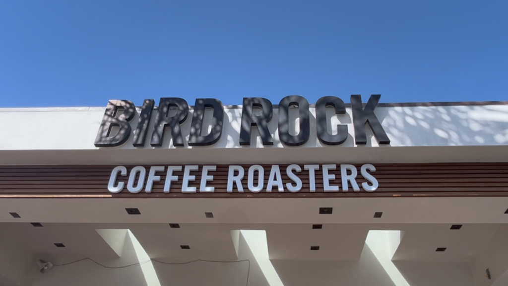 聖地牙哥 Bird Rock Coffee Roasters 咖啡廳建築外觀，深藍色木質背景配上白色品牌標誌，戶外陽光灑在寬敞的座位區長椅上