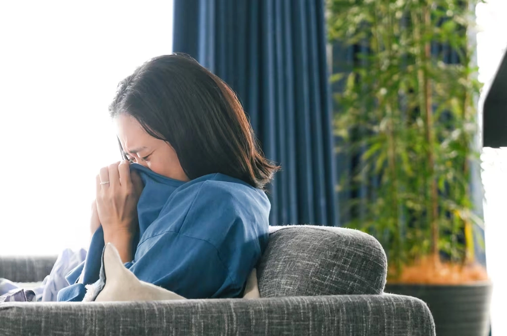 《直到 T 恤乾了為止》劇照，一名女子坐在灰色沙發上，把藍色衣服按在臉上壓抑地哭泣，背景是藍色窗簾與竹子盆栽