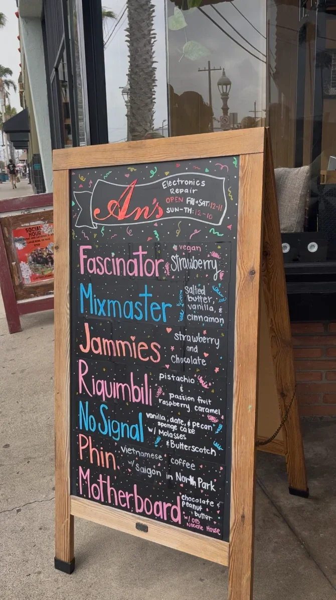 An's Electronics Repair 門口的木框 A 字黑板，用彩色粉筆寫著營業時間與當天 7 款口味：Fascinator、Mixmaster、Jammies、Riquimbili、No Signal、Phin、Motherboard，背景是 Ocean Beach 街景與棕櫚樹