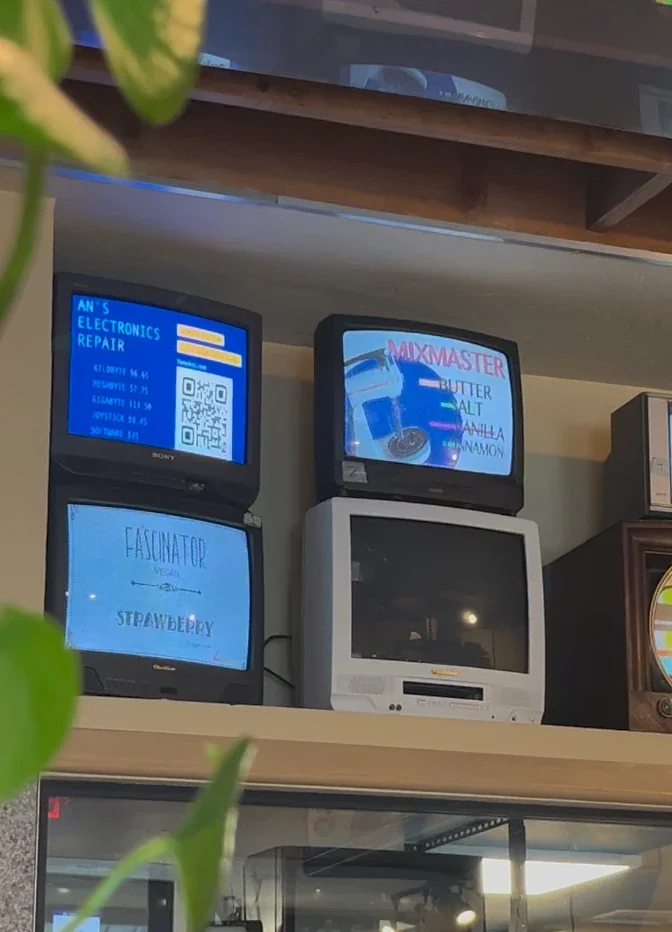 店內 CRT 老電視螢幕顯示 An's Electronics Repair 的價目表與 QR code，下方電視播放 Fascinator 草莓口味，右邊是 Mixmaster 口味，前景有綠色植物葉片