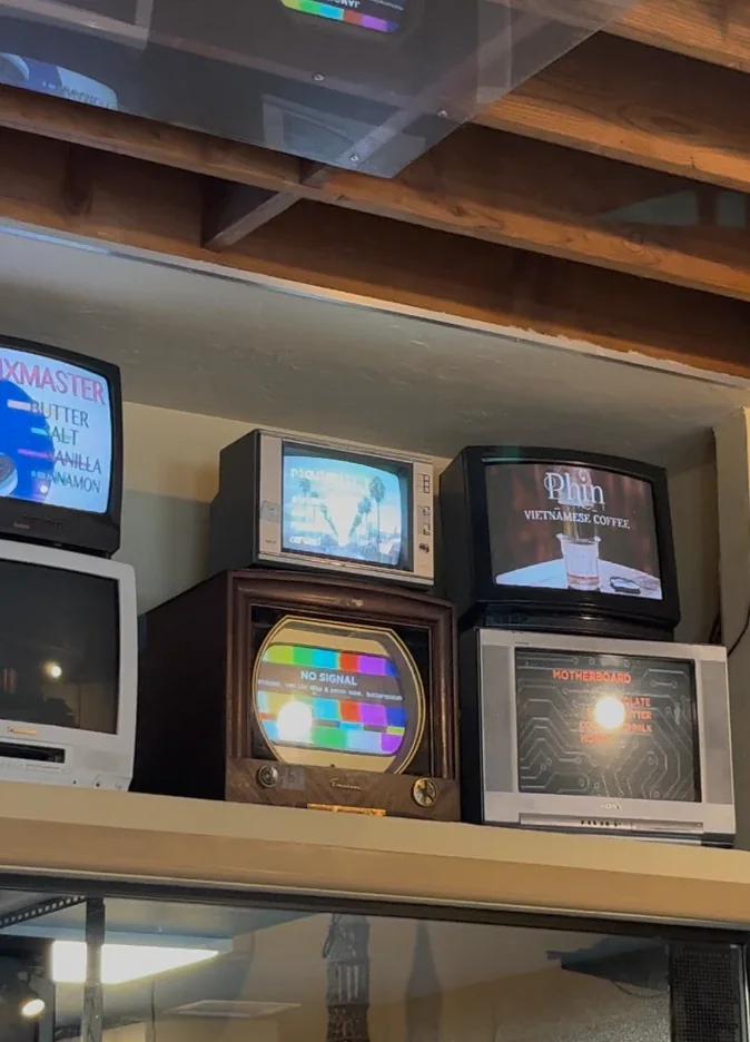 CRT 老電視牆輪播口味名稱：Mixmaster、Phin 越南咖啡、Motherboard，還有一台木殼電視播放著 No Signal 彩色色條畫面