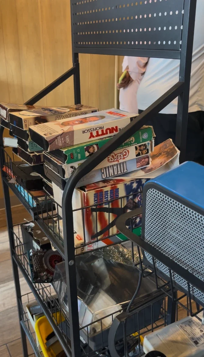 鐵架上疊著 The Nutty Professor、Grease、Titanic 等 VHS 錄影帶，旁邊有一台藍色小暖風機，背景是木質牆面