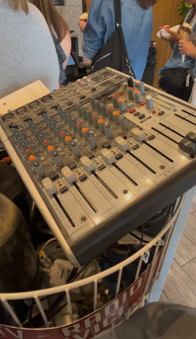 An's Electronics Repair 店內的古董混音器，擺在鐵絲籃上，旋鈕與推桿清晰可見，籃子下方印著 RADIO SERVICE 字樣，背景有排隊的客人