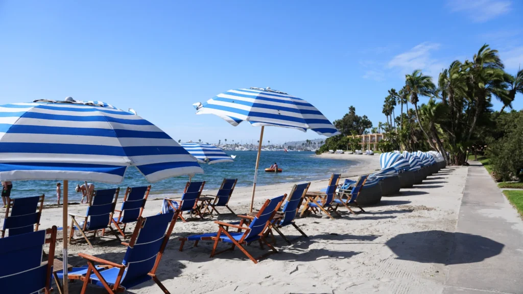 Bahia Resort Hotel 私人海灘，藍白條紋陽傘與躺椅沿著 Mission Bay 海岸排列，遠處有棕櫚樹與 cabana